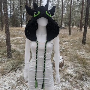 Puede incluir: Sombrero de punto negro con capucha, ojos verdes y una bufanda trenzada negra y verde a juego. El sombrero tiene forma de cabeza de dragón. "Unicorn Crochet on Etsy" está escrito en la bufanda.