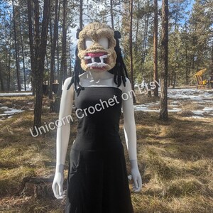 May include: A crocheted mask with a scary face, featuring white eyes, a pink mouth, and two large teeth. The mask is worn by a mannequin in a black dress, standing in a forest setting. The text "Unicorn Crochet on Etsy" is visible in the image.