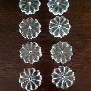 May include: Eight clear glass flower-shaped embellishments with a hole in the center. The embellishments are arranged in two rows of four.
