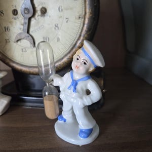May include: A vintage porcelain figurine of a sailor boy holding a glass hourglass. The figurine is dressed in a white uniform with blue accents. An antique scale is in the background.