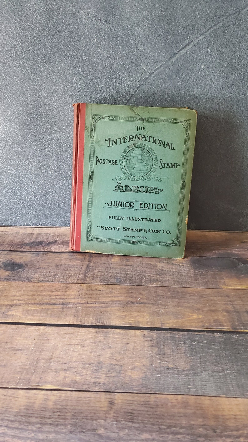 The International Postage Stamp Album Junior Edition, a green book with a globe illustration and the text "THE INTERNATIONAL POSTAGE STAMP ALBUM - JUNIOR EDITION - FULLY ILLUSTRATED - SCOTT STAMP & COIN CO. - NEW YORK"