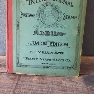 The International Postage Stamp Album Junior Edition, a green book with a globe illustration and the title in gold lettering. It is fully illustrated and published by Scott Stamp & Coin Co. in New York.