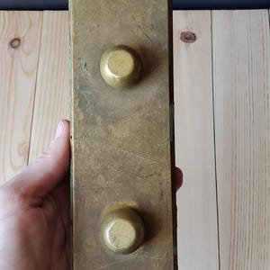 May include: A brass plate with two round, raised buttons. The plate has a worn, aged look and is slightly tarnished.