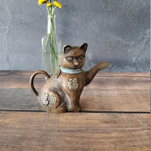 May include: A bronze cat teapot with floral details and a raised paw. The teapot is sitting on a wooden surface with a vase of yellow flowers in the background.