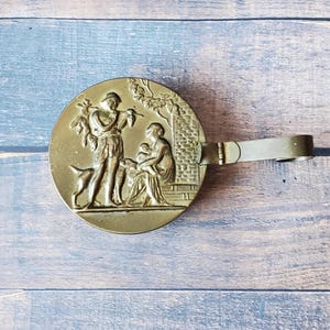 May include: A brass trinket box with a hinged lid and a handle. The lid features a relief sculpture of a man playing a flute, a woman holding a child, and a dog.