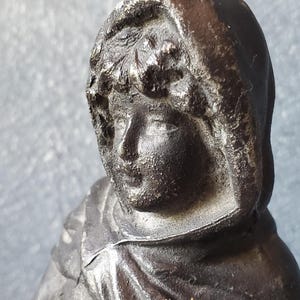 May include: A black and silver cast iron statue of a person wearing a hooded cloak. The statue is detailed with a textured surface and a realistic facial expression.
