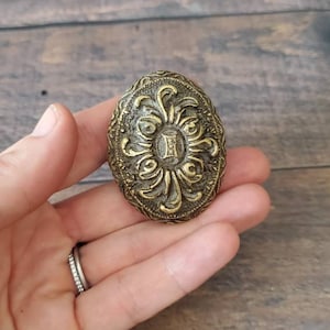 May include: A gold-toned oval-shaped metal knob with an intricate floral design and a Roman numeral II in the center.