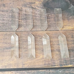 May include: Eight clear glass prism pendants with a pointed tip and a hole at the bottom. The prisms are arranged in two rows of four on a wooden surface.