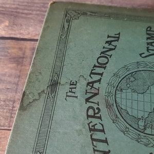 a close-up image of light water damage to the cover of the international stamp collection book . The cover of a green book with a black border and the text "The International Stamp" in black lettering. A globe is centered on the cover.
