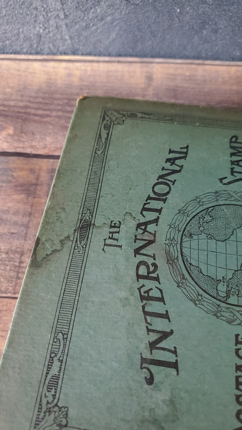 a close-up image of light water damage to the cover of the international stamp collection book . The cover of a green book with a black border and the text "The International Stamp" in black lettering. A globe is centered on the cover.