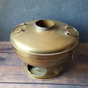 May include: A brass hot pot with a round base and a lid with two handles. The pot has a hole in the center of the base for a heat source.