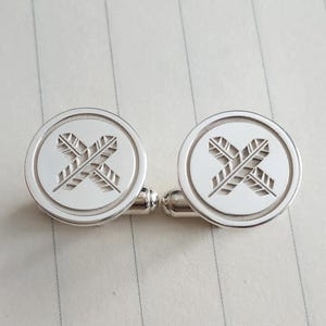 Könnte beinhalten: Ein Paar silberne Manschettenknöpfe. Jeder Manschettenknopf hat ein rundes Design mit einem gekreuzten Feder-Muster in der Mitte. Die Manschettenknöpfe liegen auf einer linierten, cremefarbenen Oberfläche.
