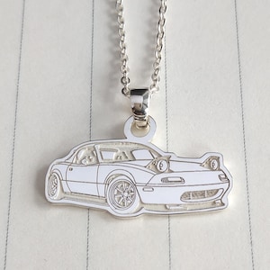 May include: A silver necklace with a pendant depicting a white sports car. The car pendant has detailed outlines of the car's features, including the wheels, headlights, and windows. The necklace is on a light-coloured, lined background.