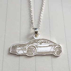 May include: A silver pendant necklace with a car design. The pendant depicts a detailed side profile of a car, suspended from a delicate chain. The necklace is shown against a light-coloured background, with the car's features clearly visible.