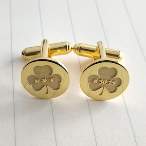 May include: A pair of gold-colored cufflinks with a clover design. One cufflink has the initials "R&T" engraved, and the other has the date "8.30.25". The cufflinks are on a white, lined surface.