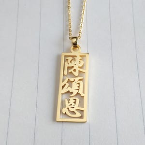 May include: A gold-coloured pendant necklace with a rectangular charm bearing Chinese characters. The charm hangs from a delicate gold chain. The pendant's design is open, allowing the characters to be seen.