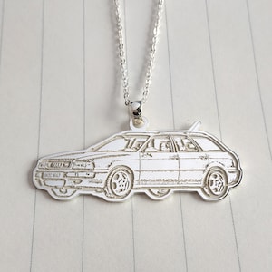 May include: A silver pendant necklace with a car design. The pendant depicts a side profile of a station wagon with etched details, suspended from a delicate chain. The car's license plate reads "INDC 647".