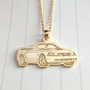 May include: Gold-tone pendant necklace with a convertible car design. The pendant is a detailed outline of the car, suspended from a delicate gold chain. The necklace is displayed on a white, lined surface.