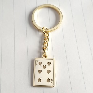 May include: Gold-colored keychain featuring a playing card design. The card is the seven of hearts, with seven heart symbols and the number seven at the top and bottom. The keychain has a gold ring and chain.