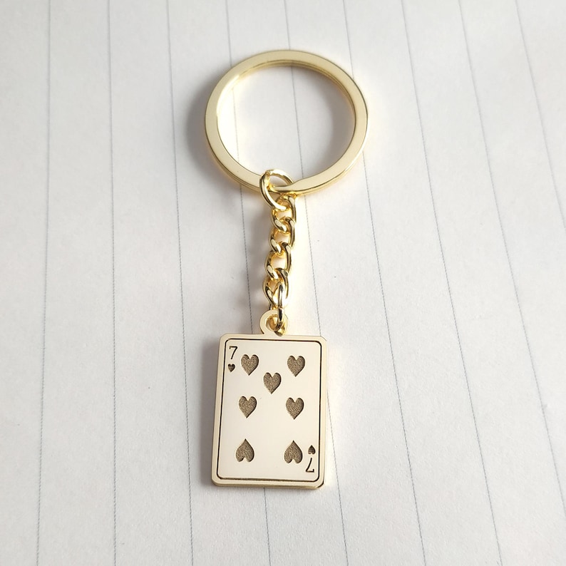 May include: Gold-colored keychain featuring a playing card design. The card is the seven of hearts, with seven heart symbols and the number seven at the top and bottom. The keychain has a gold ring and chain.