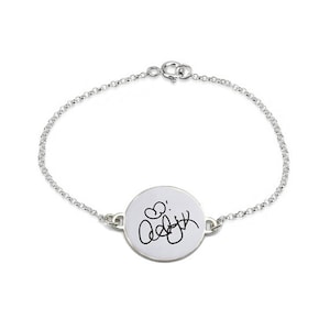 Könnte beinhalten: Ein silbernes Kettenarmband mit einem runden Anhänger, auf dem eine handschriftliche Signatur und ein Herz zu sehen sind.
