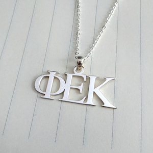 May include: Silver necklace with a pendant featuring the letters "ΦΕΚ" in a bold, blocky font.