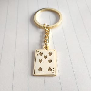 May include: Gold-colored keychain featuring a playing card design. The card is the seven of hearts, with seven heart symbols and the number seven in the corners. The keychain has a gold ring and chain.