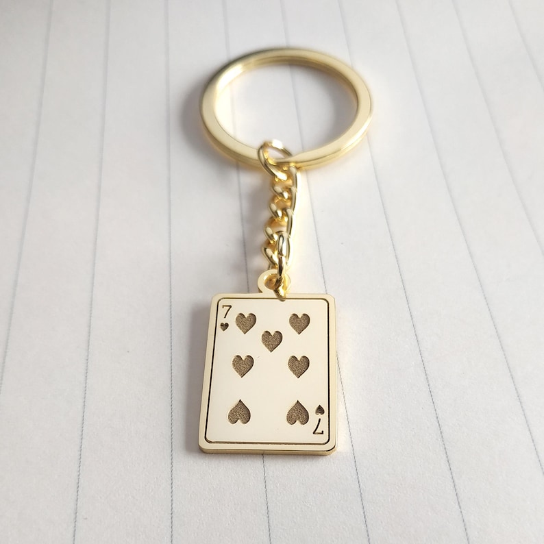 May include: Gold-colored keychain featuring a playing card design. The card is the seven of hearts, with seven heart symbols and the number seven in the corners. The keychain has a gold ring and chain.