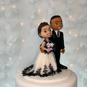 May include: A wedding cake topper featuring a bride and groom figurine. The bride is wearing a white dress with black lace trim and a bouquet of purple flowers. The groom is wearing a black suit and tie. The figurines are standing on a white cake base.