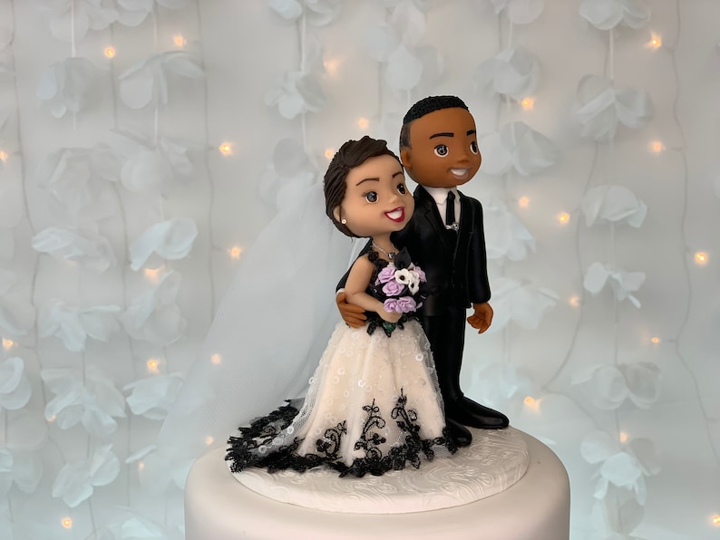 May include: A wedding cake topper featuring a bride and groom figurine. The bride is wearing a white dress with black lace trim and a bouquet of purple flowers. The groom is wearing a black suit and tie. The figurines are standing on a white cake base.