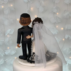 May include: A wedding cake topper featuring a bride and groom in a black tuxedo and white wedding dress with a black lace trim. The bride is wearing a white veil. The couple is facing away from the camera.
