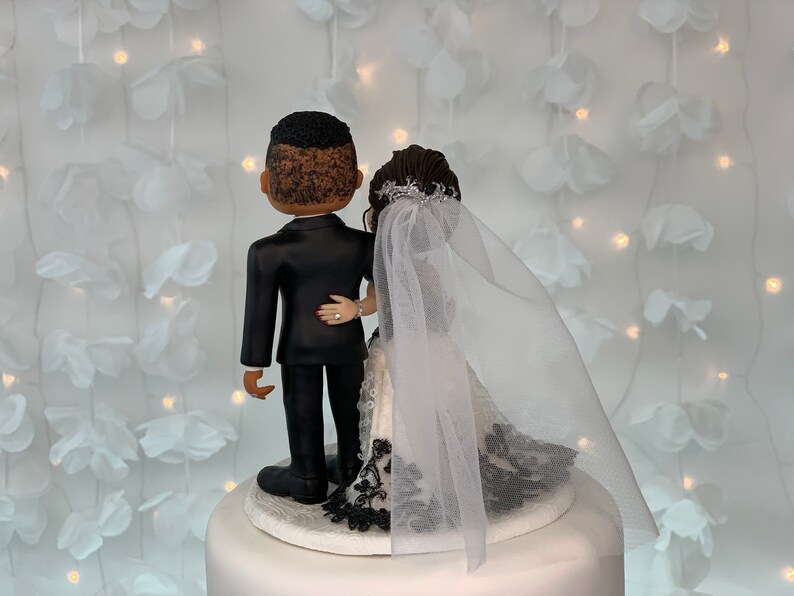 May include: A wedding cake topper featuring a bride and groom in a black tuxedo and white wedding dress with a black lace trim. The bride is wearing a white veil. The couple is facing away from the camera.