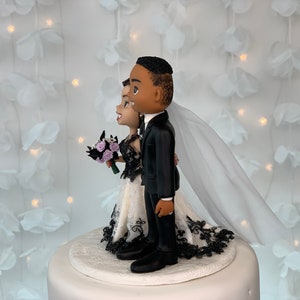 May include: Black and white wedding cake topper featuring a bride and groom in a stylized cartoon style. The bride is wearing a white dress with black lace trim and a veil. The groom is wearing a black suit. The couple is standing on a white cake base.