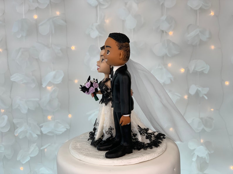 May include: Black and white wedding cake topper featuring a bride and groom in a stylized cartoon style. The bride is wearing a white dress with black lace trim and a veil. The groom is wearing a black suit. The couple is standing on a white cake base.