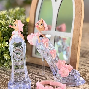 May include: Clear glass high-heeled shoe figurines with pink flower accents and mouse characters. A pink flower bracelet and a pearl necklace are also present. The background includes a wooden framed mirror and greenery.