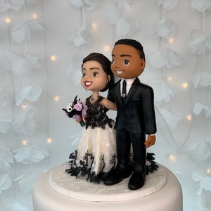 May include: A wedding cake topper featuring a bride and groom in a cartoon style. The bride is wearing a white dress with black lace and a bouquet of purple flowers. The groom is wearing a black suit and a black tie. The couple is standing on a white cake base.
