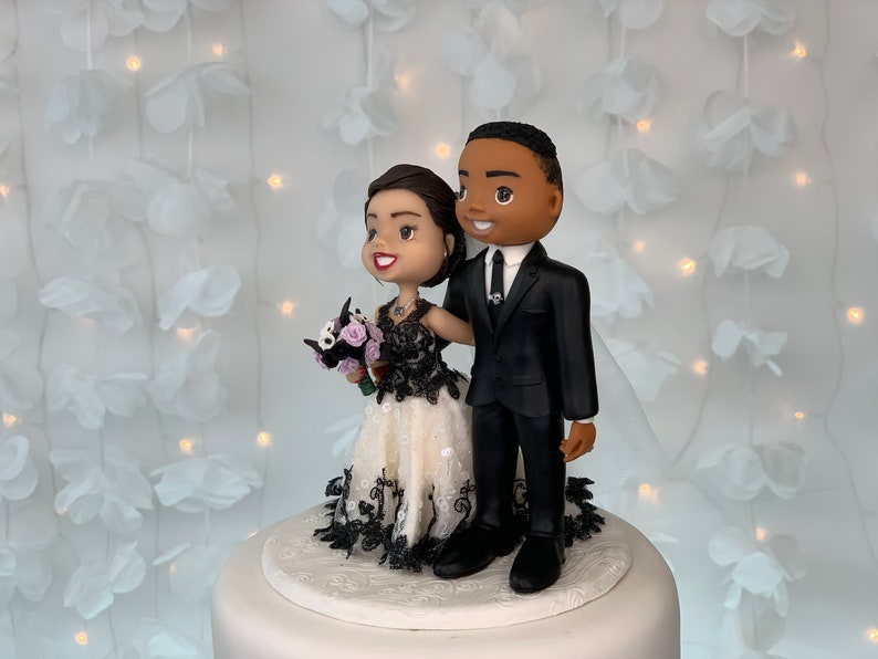 May include: A wedding cake topper featuring a bride and groom in a cartoon style. The bride is wearing a white dress with black lace and a bouquet of purple flowers. The groom is wearing a black suit and a black tie. The couple is standing on a white cake base.