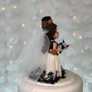 May include: A wedding cake topper featuring a bride and groom figurine. The bride is wearing a white dress with black lace trim and a veil. The groom is wearing a black suit. The couple is standing side by side, with the groom's arm around the bride's waist. They are both looking to the right.
