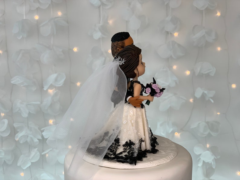 May include: A wedding cake topper featuring a bride and groom figurine. The bride is wearing a white dress with black lace trim and a veil. The groom is wearing a black suit. The couple is standing side by side, with the groom's arm around the bride's waist. They are both looking to the right.