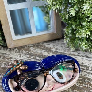 May include: A quirky ceramic eyeglass holder shaped like a face, with blue hair and a pink face. The holder features a pair of tortoiseshell-framed eyeglasses and a decorative dragonfly. A black button is also visible.