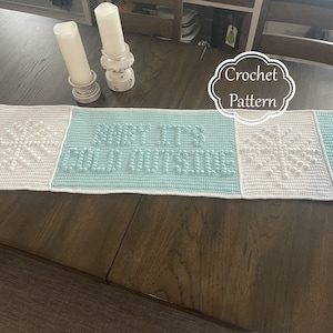 May include: Crochet table runner with a blue and white snowflake design. The runner has the text "It's Cold Outside" in the center.