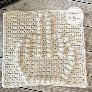 Könnte beinhalten: Cremefarbenes Häkelquadrat mit einem Kronenmuster aus erhabenen Maschen. Der Text "Crochet Pattern" befindet sich in einem weißen Kreis. Das Quadrat liegt auf einer Holzoberfläche.