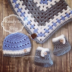 May include: A crocheted baby blanket in gray, white, and light blue with matching crocheted booties and a hat. The blanket has a square pattern and the booties and hat have a white trim.