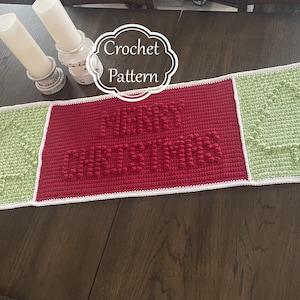 May include: A crocheted table runner with a red "Merry Christmas" design and green Christmas trees on either end. The runner has a white border and a fringe.