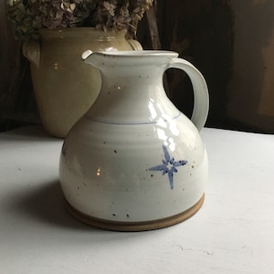 Op de afbeelding: Een witte keramische kan met een afgeronde body, een handvat en een tuit. Het heeft een blauw sterontwerp en een bruine basis. Op de achtergrond staat een tweede, beige keramische vaas met gedroogde bloemen.