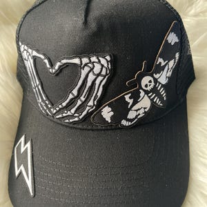 May include: Black trucker hat with a white skeleton hand making a heart shape and a white moth with a skull on its head. The hat has a black mesh back and a black lightning bolt on the front.