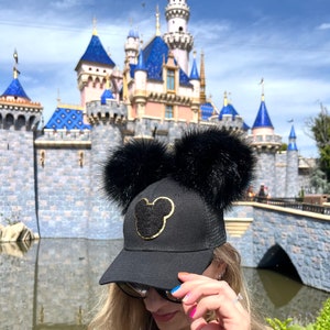 May include: A black baseball cap with black faux fur earmuffs and a gold Mickey Mouse silhouette. The cap is in front of a blue and pink castle with white towers.