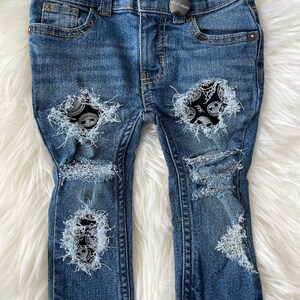 Puede incluir: Vaqueros de mezclilla azul con detalles desgastados y parches con estampado en blanco y negro. Los vaqueros tienen un ajuste skinny y cierre de botón. El nombre de la marca "Booosh" es visible en la cintura.