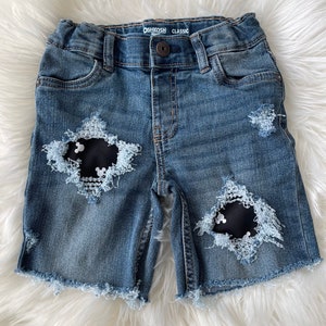 May include: Blue denim shorts with distressed details and black fabric patches featuring white Mickey Mouse silhouettes. The shorts have a frayed hem and the OshKosh B'Gosh Classic label is visible on the waistband.