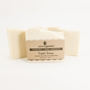 May include: Three off-white soap bars on a white background. The center bar has a brown label with the text "Fresh Snow" and "Clean & Crisp Daily Cleansing Bar". The soap is made with coconut milk and is vegan.